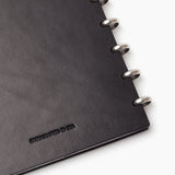 Close-up of the corner of a black Discbound Heirloom Journal (Standard 6" x 8.5") with full-grain leather and silver metal rings; HANDCRAFTED IN USA is embossed on the cover.