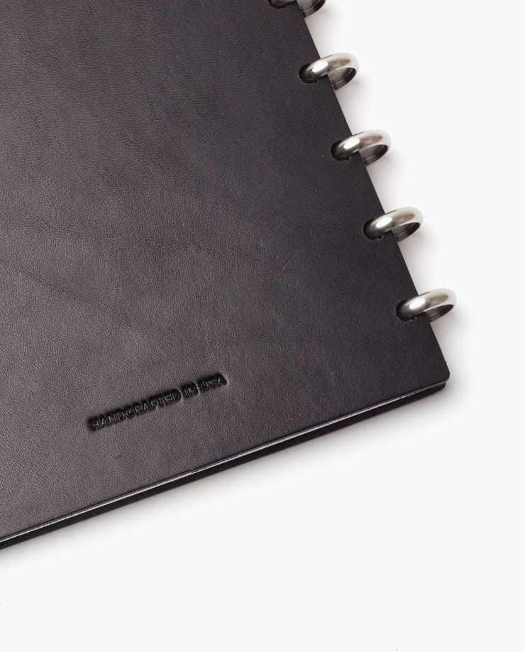 Close-up of the corner of a black Discbound Heirloom Journal (Standard 6" x 8.5") with full-grain leather and silver metal rings; HANDCRAFTED IN USA is embossed on the cover.