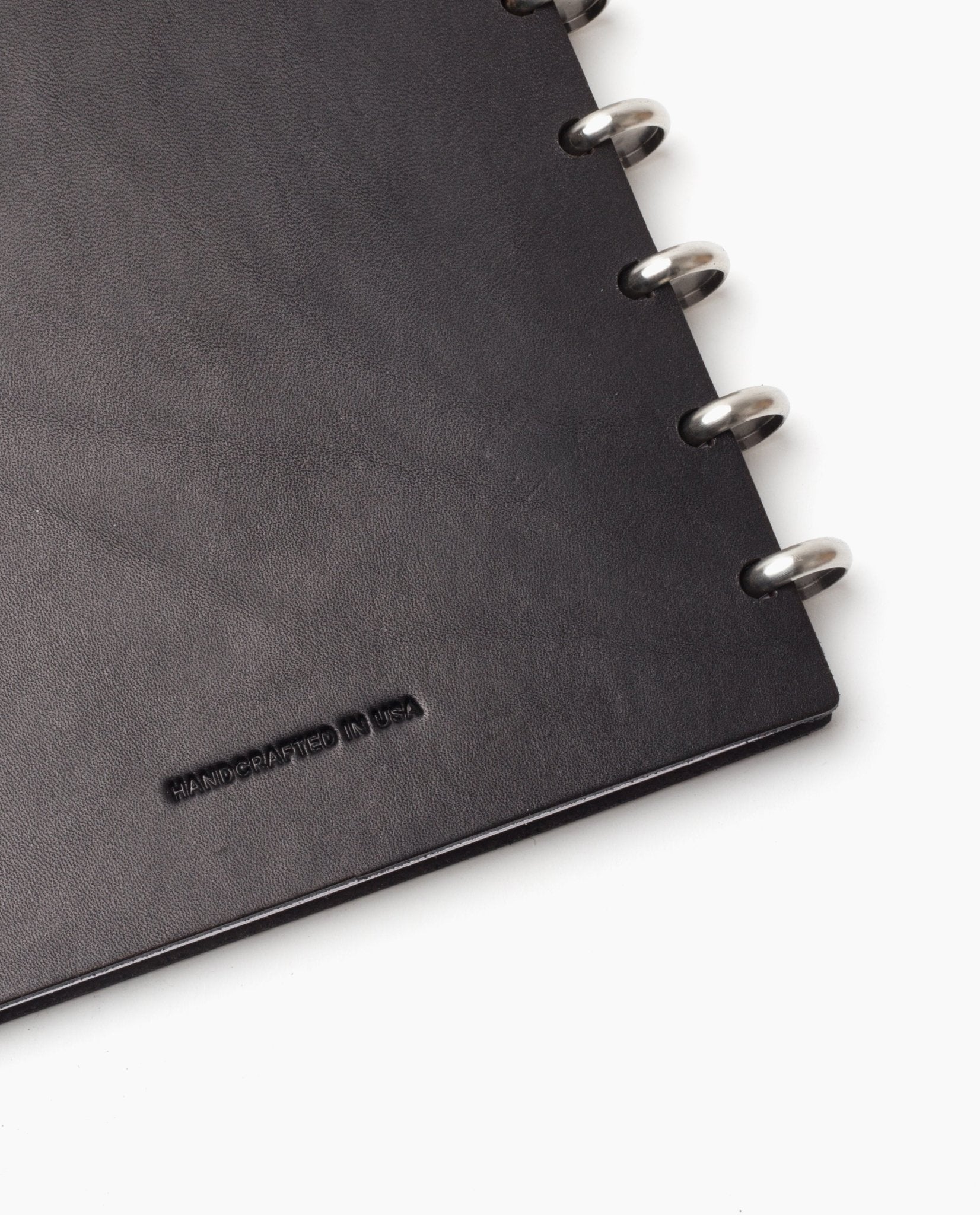 Close-up of the Discbound Heirloom Journal Bundle (Black Leather - Standard 6" x 8.5" + 3 Refills) with silver binding rings; the corner is stamped HANDCRAFTED IN USA in small black letters on a white background.