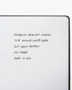 Open discbound journal with dot grid paper. Handwritten text reads: “Discbound Heirloom Journal Bundle (Tan Leather - Standard 6" x 8.5" + 3 Refills), 70# smooth white paper, dot grid pattern, 120 pages per refill, refillable notebook made in USA.”.