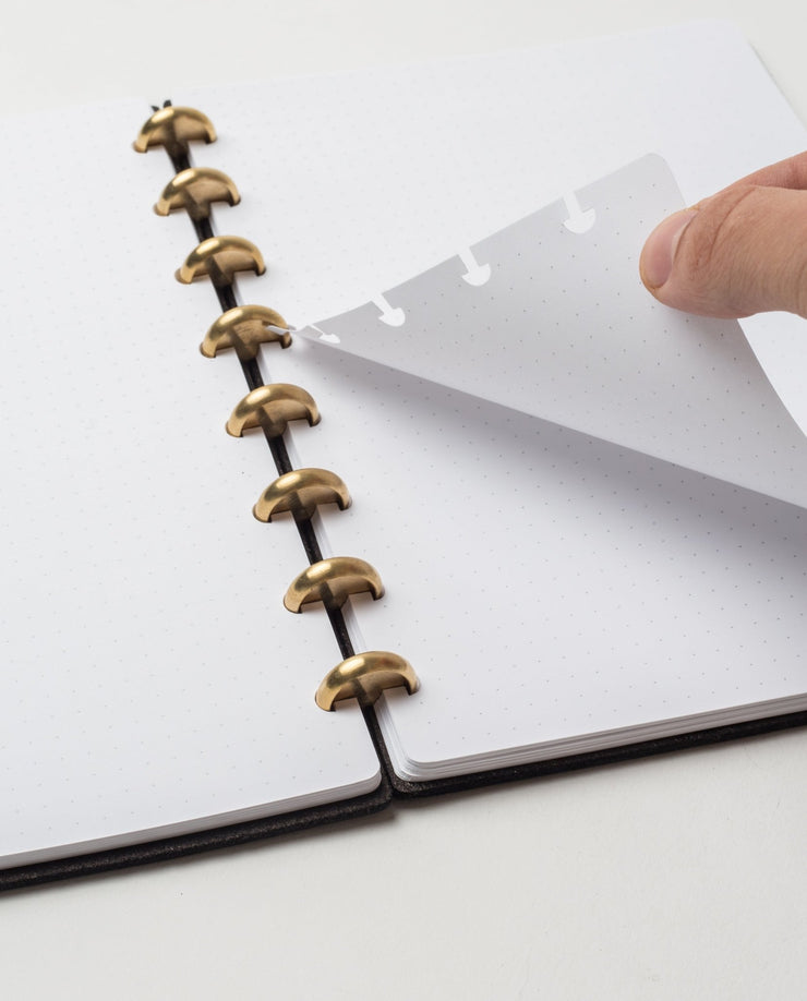 A hand lifts a dotted page in the Discbound Heirloom Journal (Black Leather - Standard 6" x 8.5"), showing its flexible, refillable design with gold disc binding and black spine. The open journal rests on a white surface.