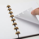 A hand lifts a dot-grid page from the Discbound Heirloom Journal Bundle (Black Leather - Standard 6" x 8.5" + 3 Refills), open on a white surface with gold disc binding and removable, repositionable pages.
