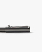 A close-up of a matte black LAMY Lx Rollerball (Ruthenium) pen with a silver clip, displayed horizontally against a white background. The image focuses on the top part of the pen, emphasizing its refillable rollerball design for smooth writing.