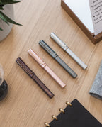 Four LAMY Lx Rollerball pens—including the elegant Ruthenium variant in dark brown, rose gold, gray, and silver—are neatly arranged on a light wooden desk near notebooks, a plant, and other stationery items. Each pen provides smooth writing performance and features a convenient refillable rollerball design for everyday use.