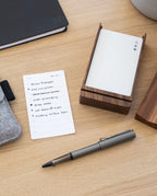 A notepad with a handwritten to-do list, pen, and wooden card holder sit on a light wood desk. Items include a notebook, felt pouch, and bowl, as well as the LAMY Lx Rollerball (Ruthenium), known for its smooth writing performance. Together, they create a neat, minimalist workspace that’s both stylish and functional.