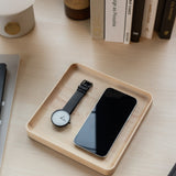 A black wristwatch and a smartphone sit side by side in a handcrafted Valet Tray (Maple - Medium) on a tidy desk, with books and other items visible in the background.