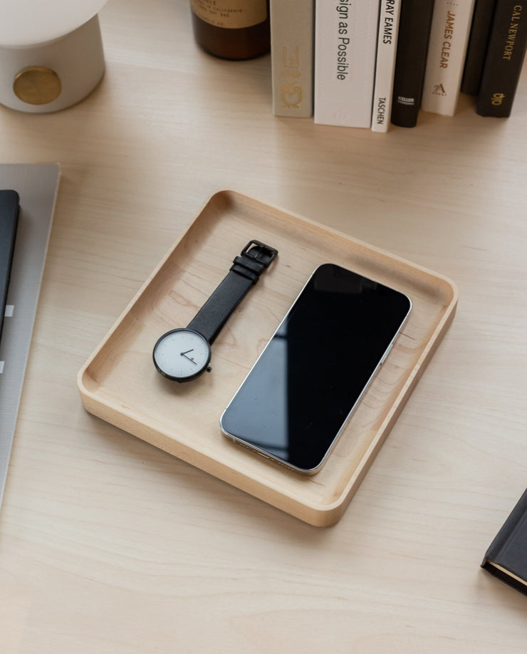 A black wristwatch and a smartphone sit side by side in a handcrafted Valet Tray (Maple - Medium) on a tidy desk, with books and other items visible in the background.