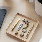 A Valet Tray (Maple - Medium) sits on a light desk, holding brass items—a pen, knife, multi-tool, keychain with keys, dice, a round object, and a small cylinder. Nearby are a plant pot and some notebooks.