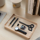 A Valet Tray Bundle (Maple - Set of 3), handcrafted in Pennsylvania, sits on a light wood desk holding pens, a watch, scissors, an eraser, a gold binder clip, and a letter U tile. Books and a lamp are arranged behind the trays.
