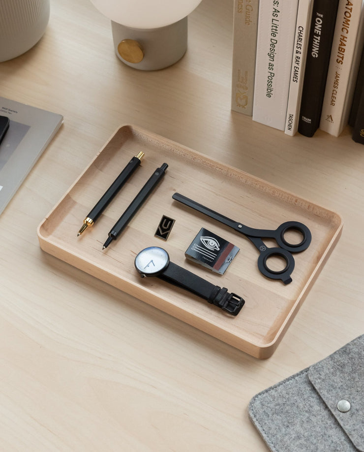 A Valet Tray Bundle (Maple - Set of 3), handcrafted in Pennsylvania, sits on a light wood desk holding pens, a watch, scissors, an eraser, a gold binder clip, and a letter U tile. Books and a lamp are arranged behind the trays.