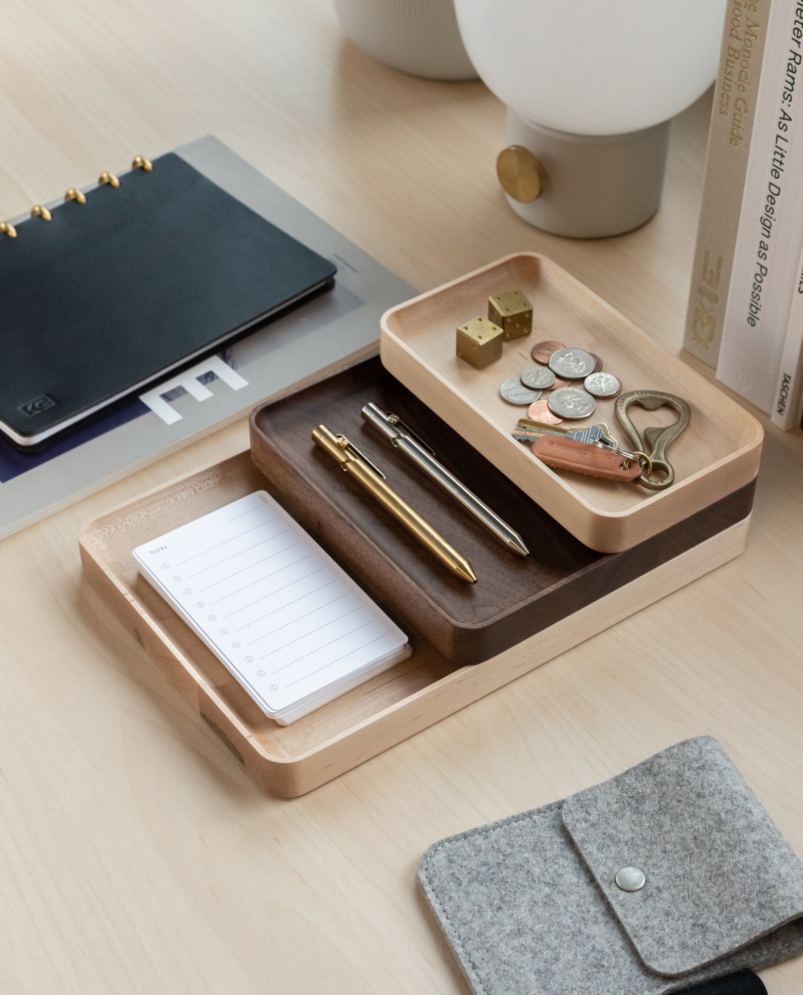 The Valet Tray Bundle (Maple - Set of 3), handcrafted in Pennsylvania, keeps pens, coins, keys, small locks, and a notepad organized on a light wood desk alongside a grey pouch, black notebook, books, and a white lamp.