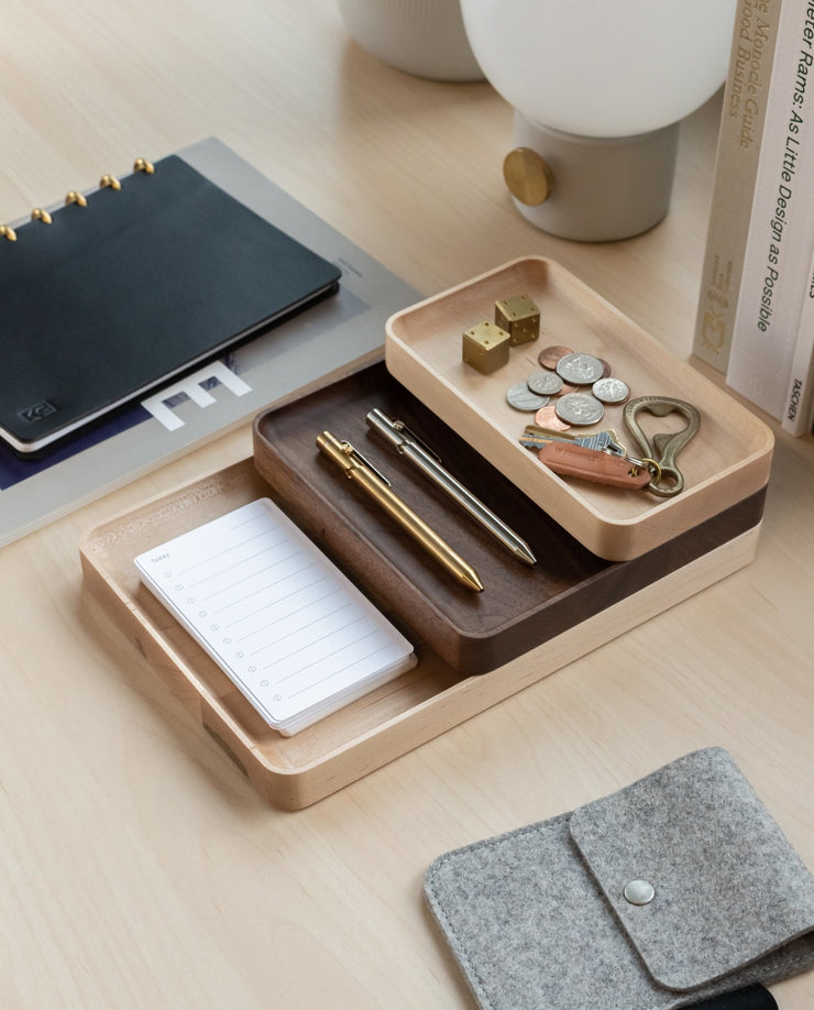 The Valet Tray Bundle (Maple - Set of 3), handcrafted in Pennsylvania, keeps pens, coins, keys, small locks, and a notepad organized on a light wood desk alongside a grey pouch, black notebook, books, and a white lamp.