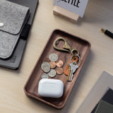 The Valet Tray (Walnut - Small), handcrafted in Pennsylvania, sits on a desk holding coins, a brass keychain bottle opener, two keys, and a white wireless earbud case beside a pen, notebooks, and a gray felt pouch for everyday carry.