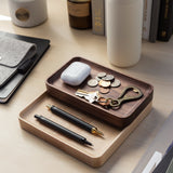 The Valet Tray (Walnut - Small), handcrafted in Pennsylvania, holds pens, keys, coins, and earbuds. Nearby sit a gray felt pouch, books, bottles, and a white tumbler on a light wood desk for everyday carry storage.