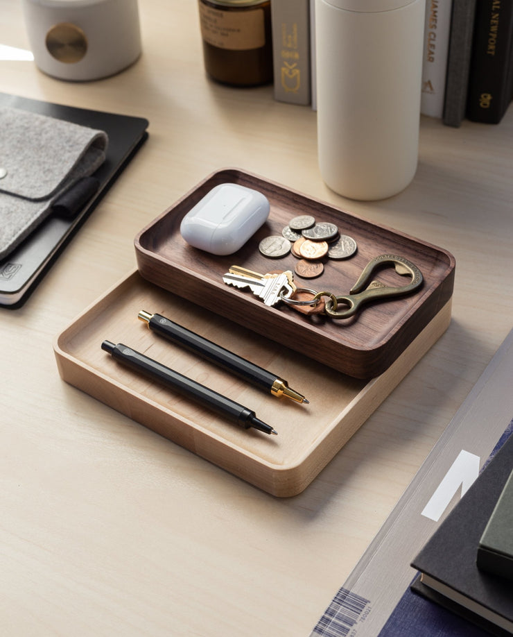 The Valet Tray (Walnut - Small), handcrafted in Pennsylvania, holds pens, keys, coins, and earbuds. Nearby sit a gray felt pouch, books, bottles, and a white tumbler on a light wood desk for everyday carry storage.