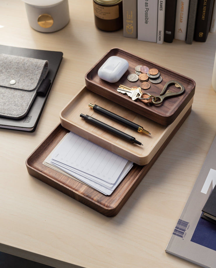 The Valet Tray (Maple - Medium) features three compartments to neatly organize coins, keys, a bottle opener, wireless earbuds, two black pens, and notepads on a light wood desk next to books and office essentials.
