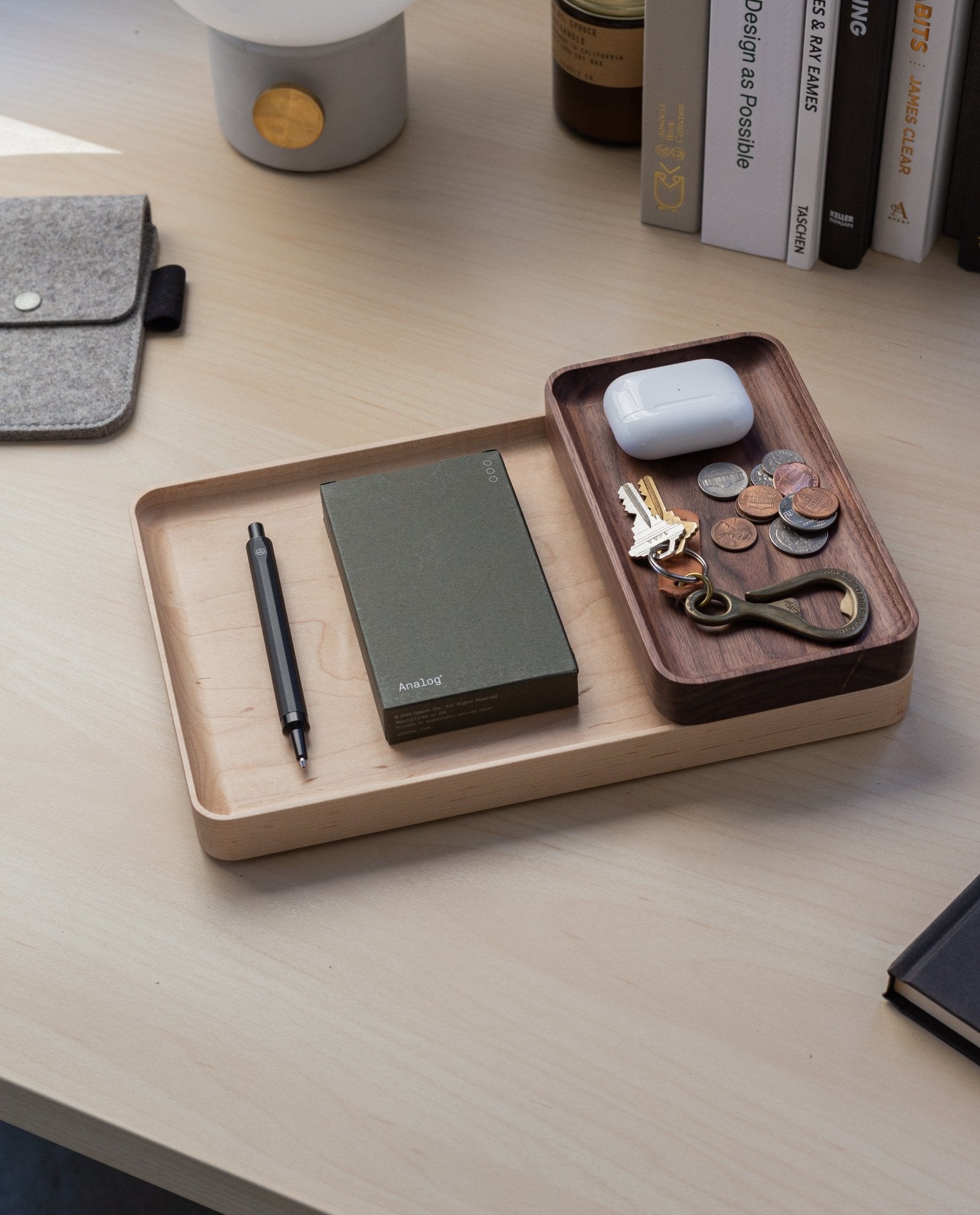 The Valet Tray Bundle (Maple - Set of 3), handcrafted in Pennsylvania, neatly holds a pen, green notebook, keys, coins, a brass bottle opener, and white wireless earbuds case on a light wood desk beside books and a gray fabric pouch.