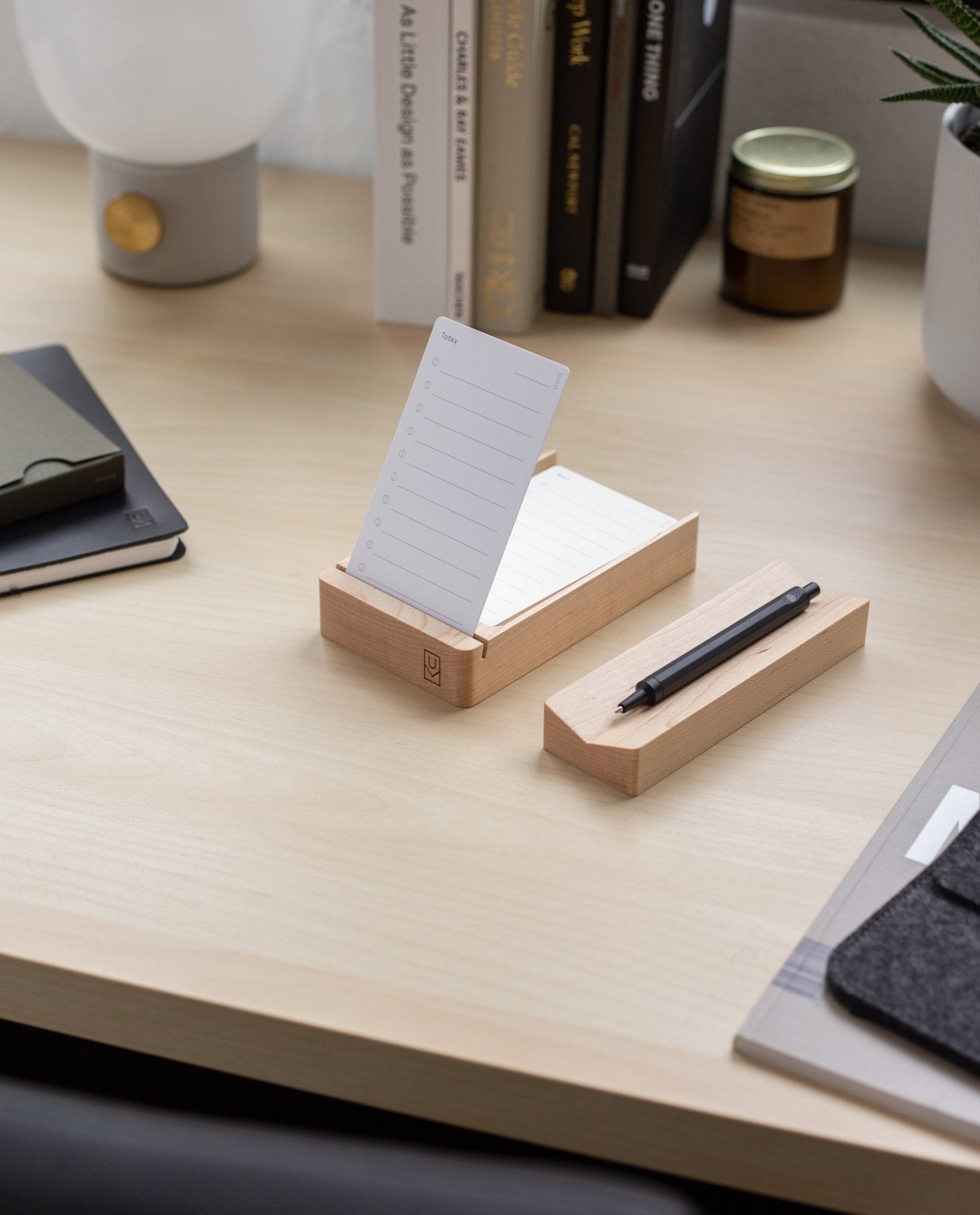A neat desk with a solid maple notecard holder displaying a lined note, the Pen Tray (Maple) holding a pen and made in Pennsylvania, books, a small plant, a lamp, a notebook, and a candle arranged on a light wood surface.