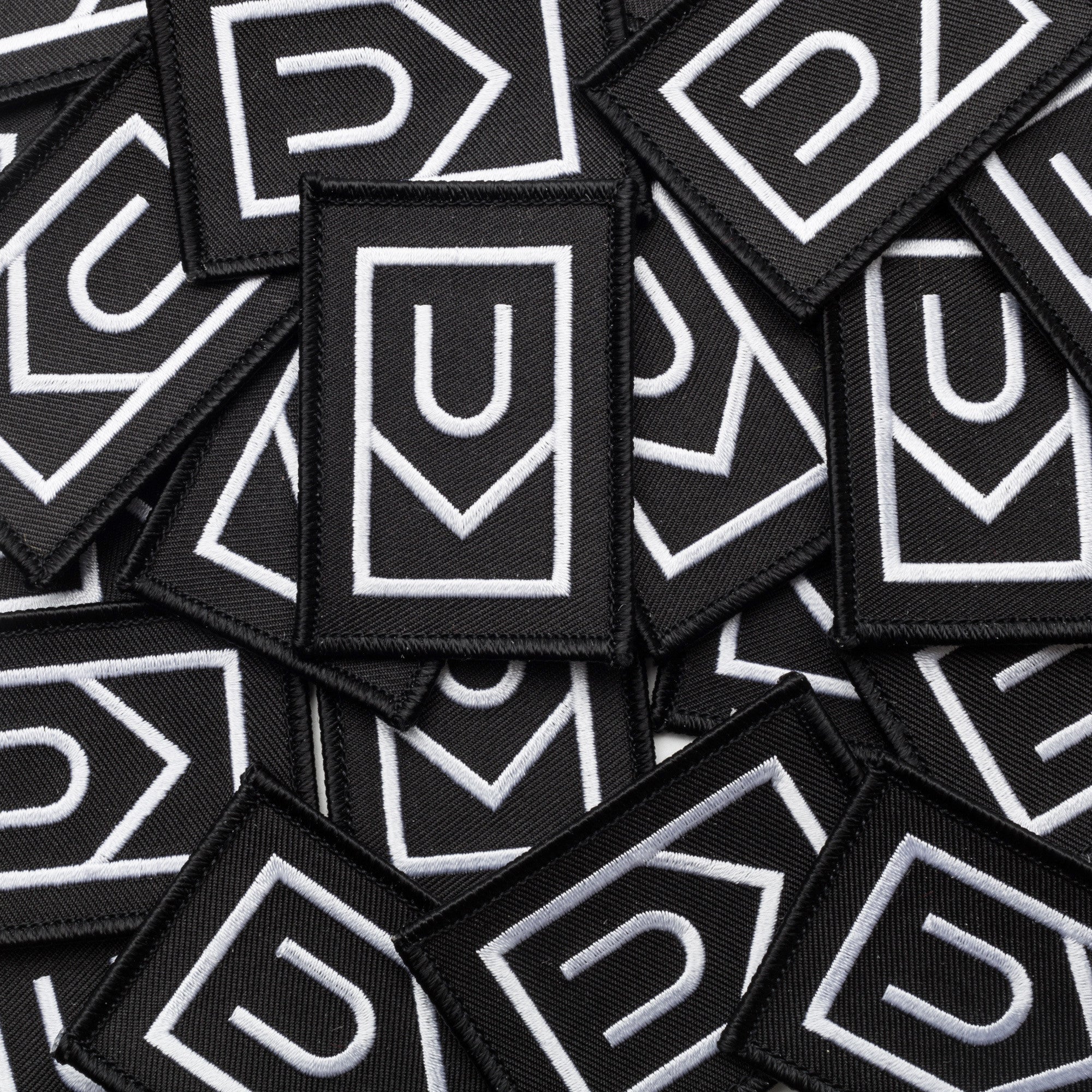 A pile of Ugmonk Patches—black, rectangular embroidered patches featuring white geometric designs with a U shape above a downward-pointing arrow.
