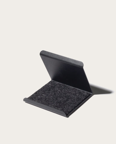 The Gather Phone Stand (1x1 - Black) is a minimalist, black, rectangular stand with a hinged lid, partially open to show its soft dark felt interior, set against a plain light background—ideal as a tablet or phone stand.