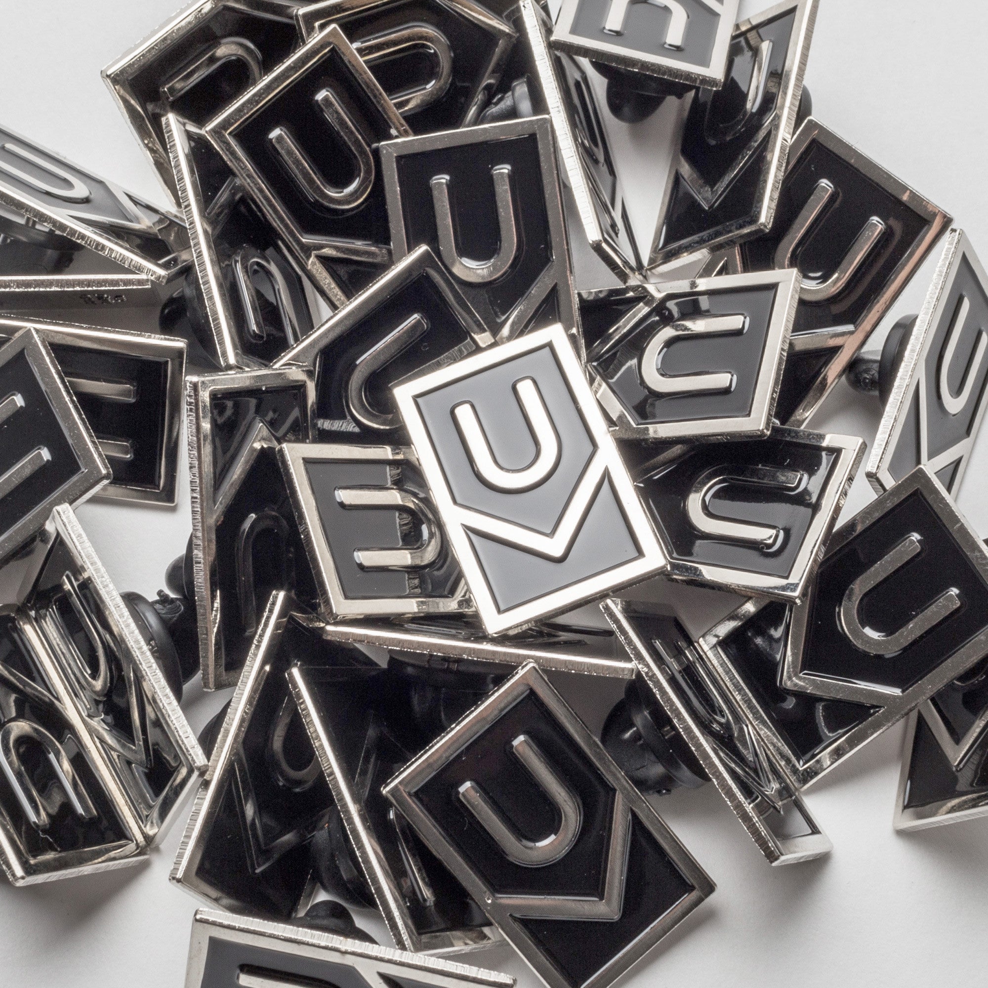 A collection of Ugmonk Enamel Pins with a geometric U-shaped design in black and silver, scattered and overlapping on a white surface.