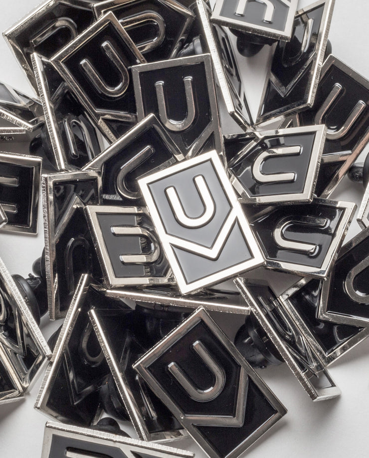 A collection of Ugmonk Enamel Pins with a geometric U-shaped design in black and silver, scattered and overlapping on a white surface.