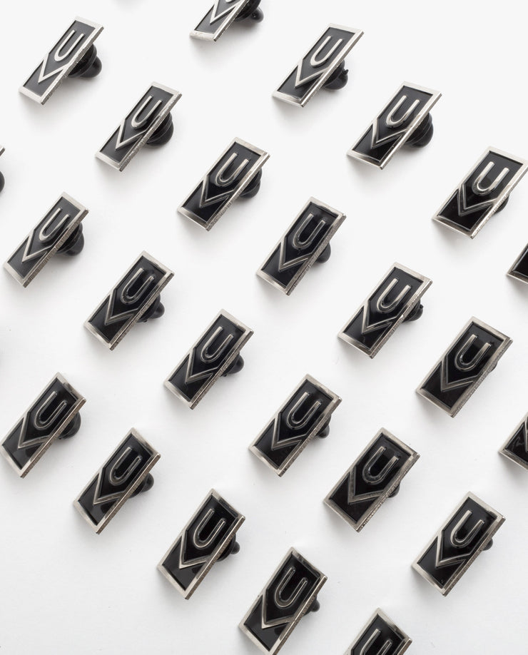 Ugmonk Enamel Pins in black rectangles and silver logos are arranged diagonally in a grid on a white background. Each pin displays a geometric U or V design.