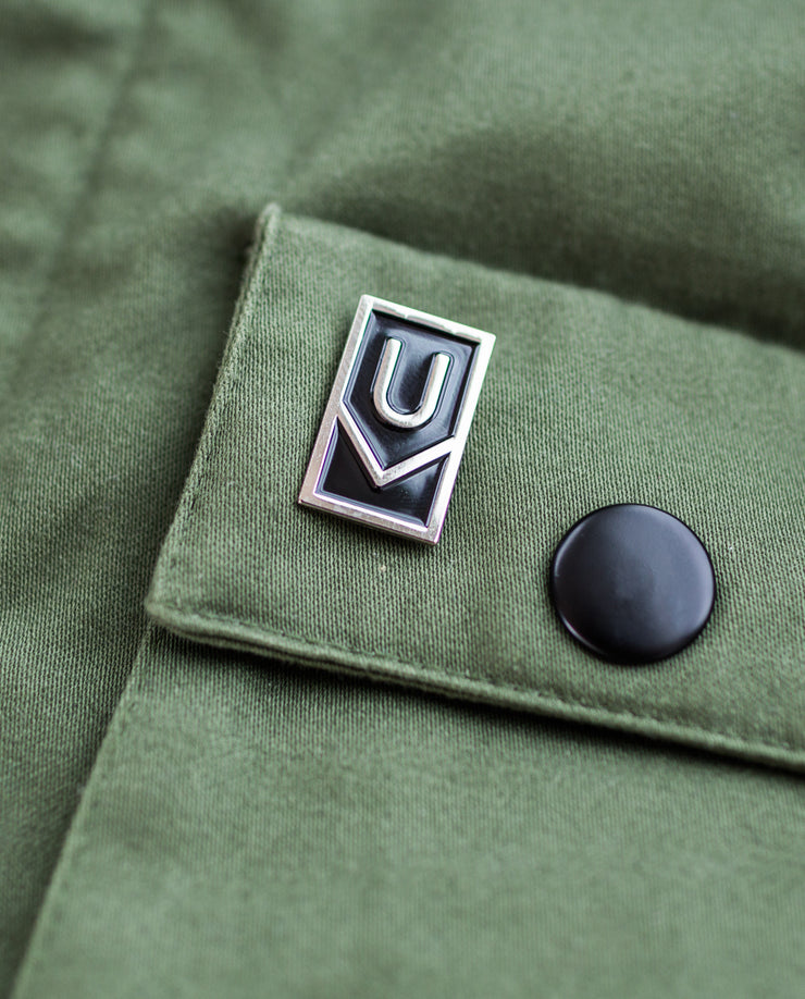 A close-up of olive green fabric featuring a black snap button and the Ugmonk Enamel Pin—a rectangular silver logo with a geometric design displaying the letters U and V.