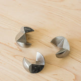 Three stainless steel geometric pieces from the Craighill Venn Puzzle, featuring crescent and triangular shapes, are arranged in a loose triangle on a light wood surface.