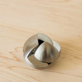 The Craighill Venn Puzzle (Stainless Steel) with interlocking, curved pieces rests on a light wood surface. This geometric, spherical mechanical puzzle has a smooth, shiny finish and is crafted from stainless steel.
