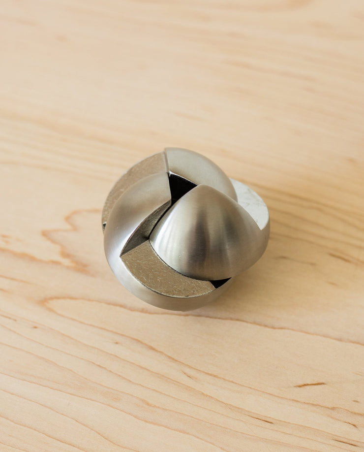 The Craighill Venn Puzzle (Stainless Steel) with interlocking, curved pieces rests on a light wood surface. This geometric, spherical mechanical puzzle has a smooth, shiny finish and is crafted from stainless steel.