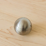 The Craighill Venn Puzzle (Stainless Steel) is a metallic, spherical puzzle with smooth, brushed surfaces and visible interlocking seams, resting on a light wooden surface.